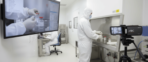 Lab technician in full cleanroom suit handling vials inside a sterile hood, with camera recording and large monitor showing close-up of the procedure. 