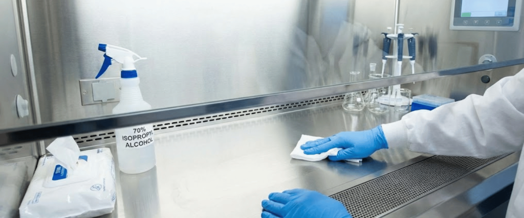 Gloved hands wiping a stainless steel laminar flow hood surface with disinfectant wipes and a bottle labeled 70% isopropyl alcohol