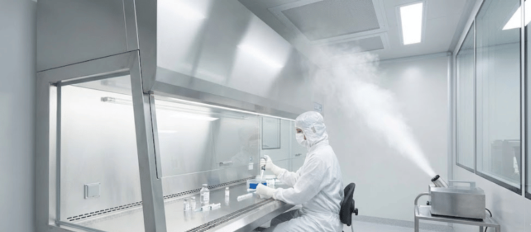 Cleanroom technician in full protective suit working at a sterile laminar flow bench, handling vials and pipettes amid fogging sterilization