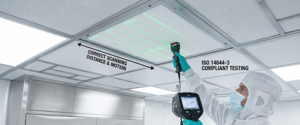 Technician in cleanroom suit and mask scanning a ceiling panel with a handheld ISO 14644-3 particle counter, green laser grid visible