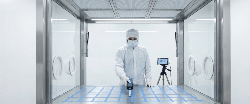 Technician in full cleanroom suit uses testing device on taped grid inside stainless steel containment with monitoring equipment