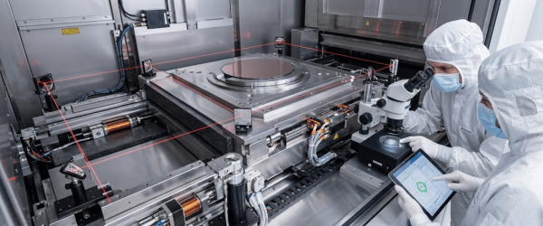 Technicians in cleanroom suits inspect a semiconductor wafer using a microscope and tablet beside precision lithography equipment