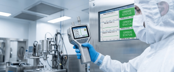 Cleanroom technician in full gown holding particle counter with monitoring panel showing green status in sterile lab environment