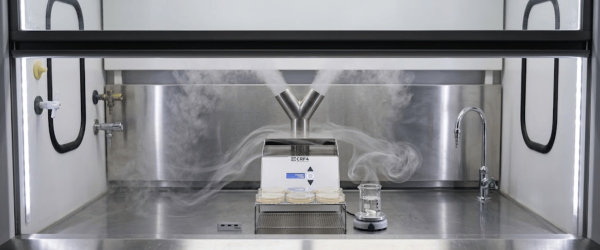 Fogger producing sterile vapor over petri dishes and glassware on a stainless steel lab bench inside a fume hood, fogger visible front-center