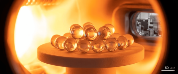 Glass microbeads stacked pyramid-style on a heated ceramic pedestal inside an orange-glowing furnace chamber, laboratory equipment visible through a window