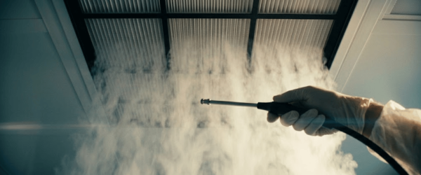 Smoke studies monitoring technician sprays smoke toward a ceiling vent to test airflow and ventilation performance, gloved hand holding nozzle