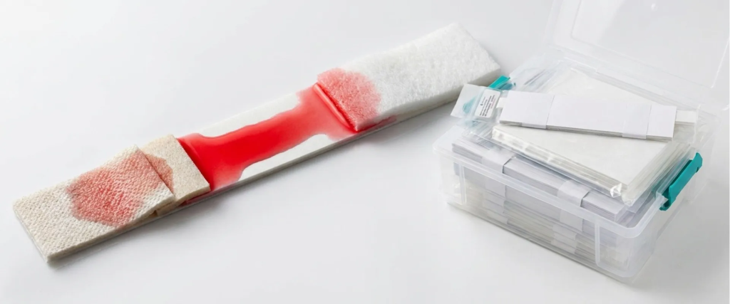 Lateral Flow test strip showing red sample spread beside a clear storage box with test materials on a white background
