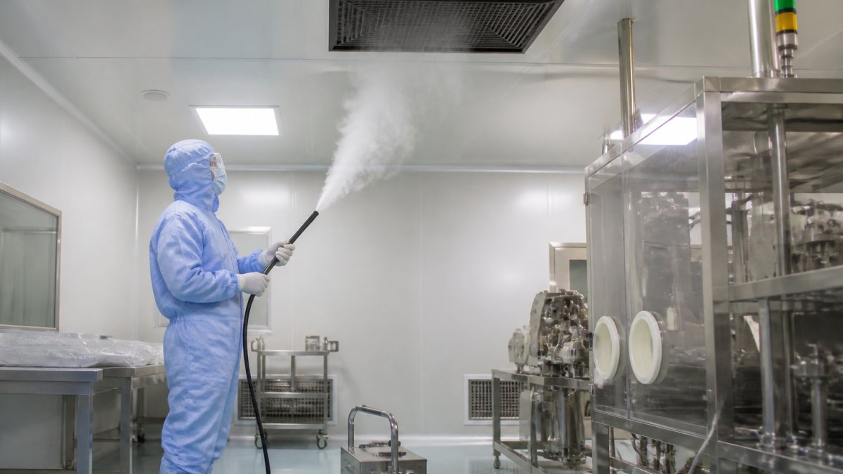 A technician in full cleanroom gowning holding a fogger wand, releasing visible white smoke to visualize unidirectional airflow from a ceiling HEPA filter in a pharmaceutical cleanroom