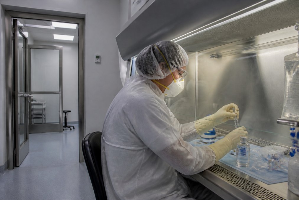 A cleanroom technician in full PPE gowning, including chemotherapy gloves and a respirator, working inside a biological safety cabinet in a negative pressure compounding suite with visible ante room behind